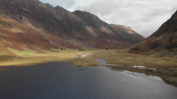 Slowly pulling back along a dark lake to reveal a Scottish highland valley in Glencoe, Scotland. Aer