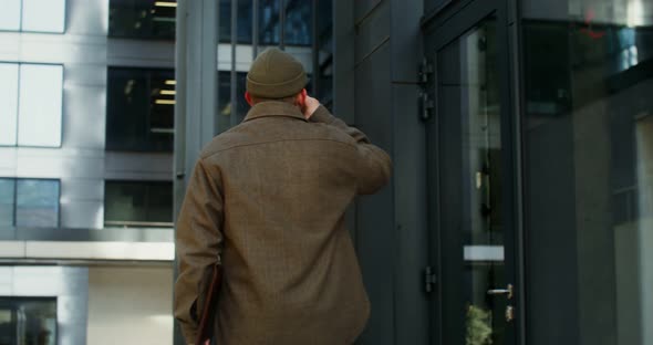 A Bearded Man Walks Near an Office Building and Drinks Coffee
