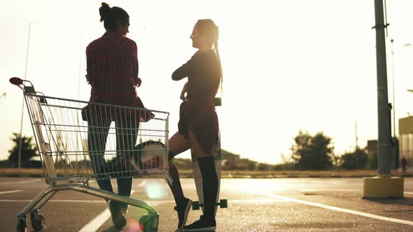 Back View of Two Attractive Stylish Girls Standing By the Shopping Cart on Parking and Talking