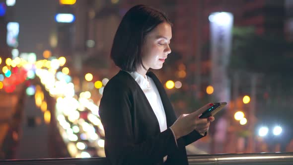 Asian young business woman standing outdoor using mobile phone downtown in city at night.