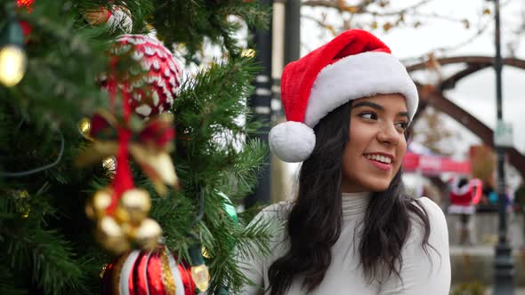A hispanic woman in a Santa Claus hat looking happy and merry next to a holiday Christmas tree with