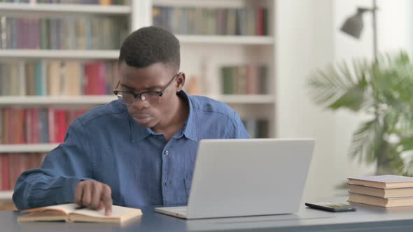 Young African Man Reading Book and Working on Laptop in Office