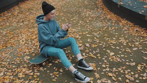 Young Caucasian Woman with Black Winter Hat Playing Games n Smartphone While Sitting on Skateboard