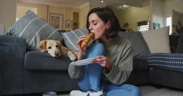 Smiling caucasian woman eating croissant and stroking her pet dog on sofa next to her