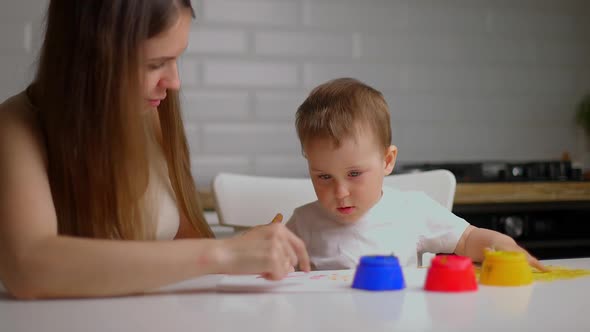 Beautiful Mom Helps Her Son to Paint with Hands Sitting at the Table in the Kitchen