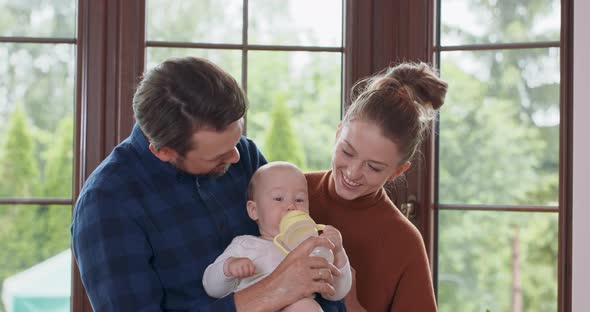 Young Happy Parents are Feeding Their Adorable Baby in Front of the Window Bearded Father Holds His