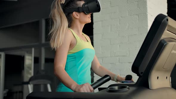 Beautiful Caucasian Girl in VR Helmet Walks on Treadmill in Gym