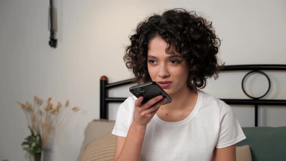 Smiling Hispanic Curly Woman Uses Smartphone Dictating Audio Message to Friend