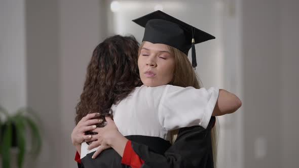 Young Female Graduate in Gown and Hat Hugging Woman with Dissatisfied Irritated Facial Expression