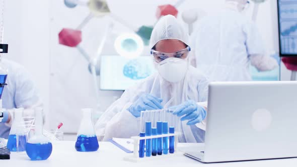 Female Scientist Picking Up a Test Tube with Blue Solution and Typing on Computer alt