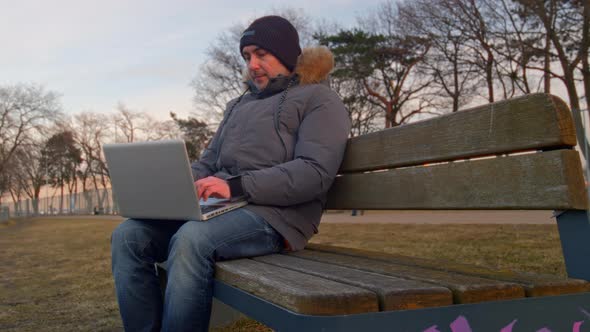 Man in the Park on a Bench Working on a Laptop in a Winter Down Jacket