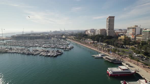 Aerial backwards view over marina with boats tied up. Alongside a busy road with trees and modern bu