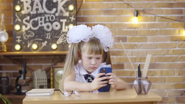 Little Girl Playing a Game on Smartphone Sitting at Table on Background of a School Class