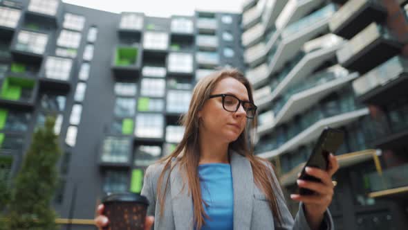 Formally Dressed Woman Walking Down the Street in a Business District with Coffee in Hand and Using