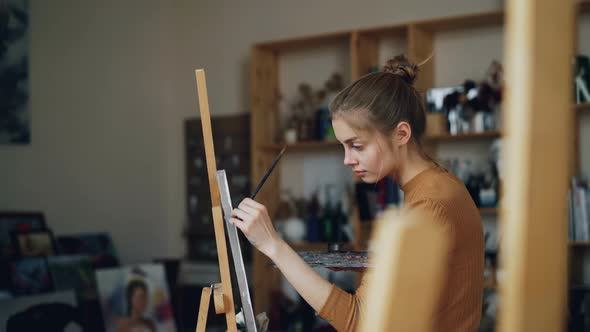 Slender Girl Artist Is Working in Studio Painting on Canvas Holding Brush in Dirty Hand and Mixing