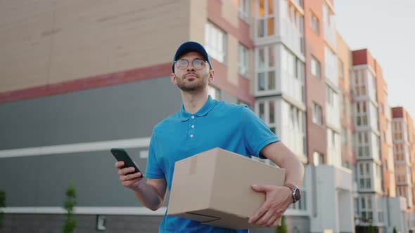 The Postman with Glasses Carries the Parcel and Looks at the Delivery Address Via Mobile Phone