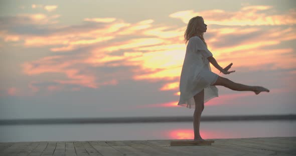 Flexible Young Lady in White Dress is Dancing on the Pier of the Lake