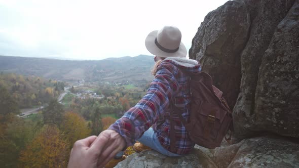 POV First Person View of Man Stretching Hand to Pretty Woman Hiker in Hat with Backpack Sitting on