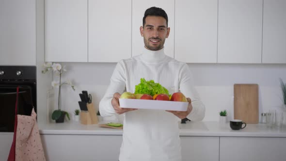 Camera Approaches to Young Positive Middle Eastern Chef Posing with Healthful Fruits and Greenery in