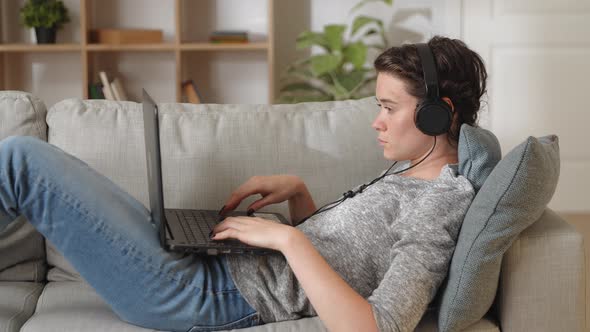 Young Serious Focused Woman Lying on Cozy Sofa Wear Wireless Headphones Studying or Working Using