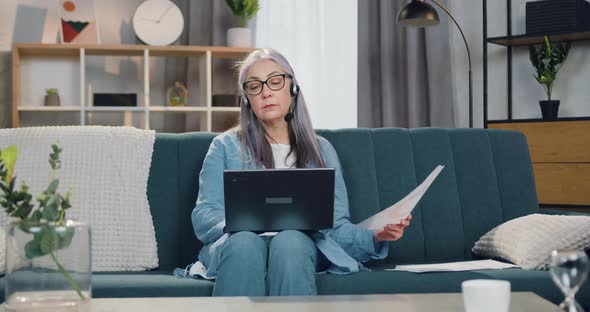 Serious confident grey-haired 65-aged woman in headset sitting on the couch and working on laptop