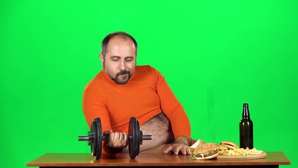 Caucasian Man Sits at a Table with Junk Food in One Hand Holds Dumbbell in Second Bottle of Beer