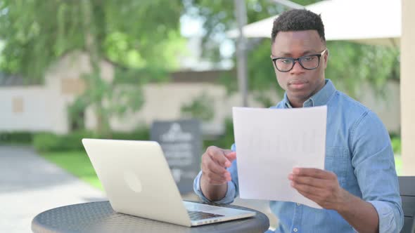 African Man with Laptop Reading Documents in Outdoor Cafe