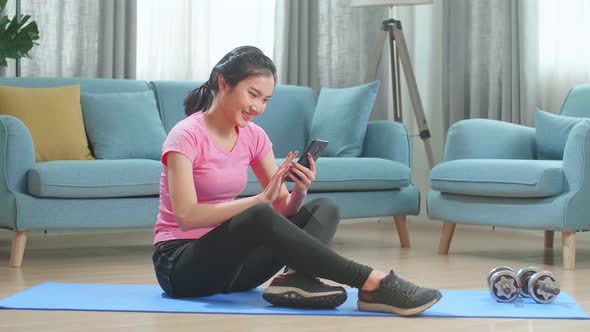 Asian Female Fitness Girl Is Using An App On Her Smartphone Before Working Out In Living Room