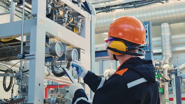 An Engineer Technician in a Hard Hat Goggles and Headphones Writes the Data From a Pressure Gauge