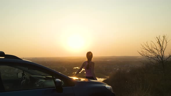 Young woman standing near her car enjoying warm sunset view. Girl traveler leaning on vehicle