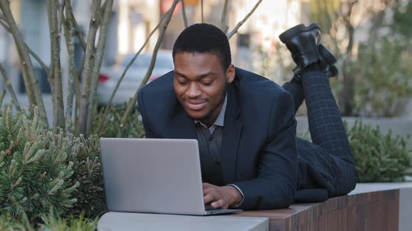 Young African American Businessman Typing on Laptop Laying on Her Stomach Outdoors Working Distantly