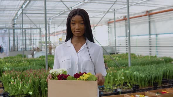 Beautiful Young African American Girl in a White Shirt Stands in a Greenhouse and Shows Off Colorful