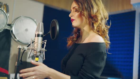 Portrait of a happy female wearing black singing into a modern microphone in audio studio.
