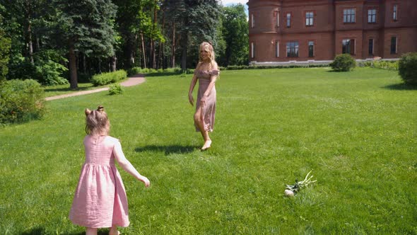 Woman and a Child in Pink Dresses Run Across a Green Field Against the Background of an Old Castle