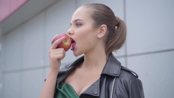 Portrait of a Beautiful Girl in an Emerald Evening Dress and a Leather Jacket Biting a Juicy Apple