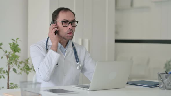 Middle Aged Doctor Talking on Smartphone While Working on Laptop