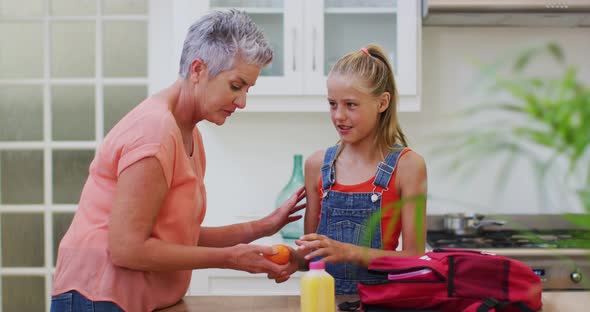 Caucasian grandmother in kitchen preparing packed lunch talking with granddaughter giving her fruit