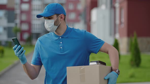 The Male Deliveryman in a Cap and a Protective Mask and Gloves Goes with a Box in His Hands and