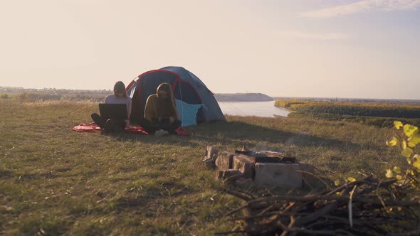 Girls with Tablet and Laptop Sit at Tent in Camp at River