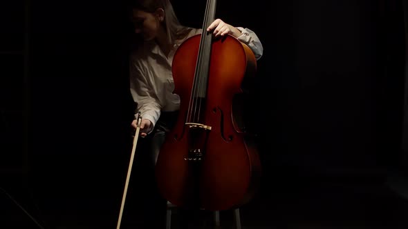 Female Musician Sits on Chair and Starts Playing Cello Front View Studio Black Background