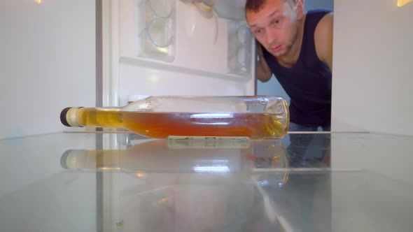 man looks into an empty refrigerator and takes out bottle of alcohol. Alcoholism