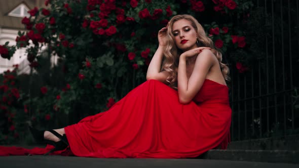 Fabulous Girl with Red Lips in Red Dress on Awesome Summer Roses Background. Fantasy Woman Portrait