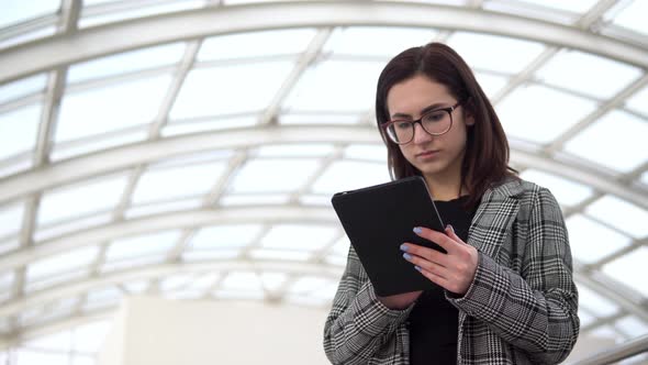A Young Woman Is Standing with a Tablet. A Girl Uses a Tablet Against a Pan-glass Roof. A Busy