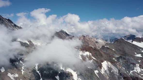 Aerial dolly out of a montain range above the clouds in a sunny summer day with snow patches