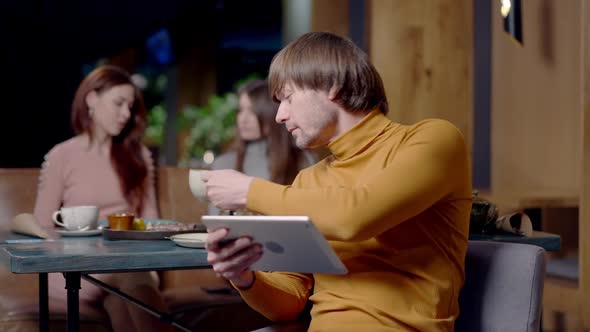 Portrait of Busy Confident Young Man Using Tablet with Blurred Women Talking at Background Sitting