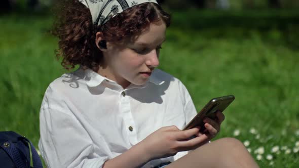 Teenage Girl is Sitting on the Green Grass in the Park and Talking to Someone on Her Cell Phone