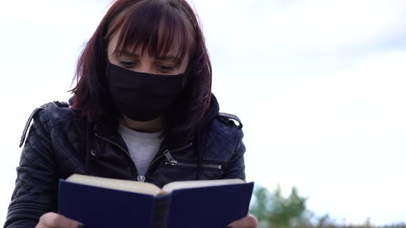 Close Up of Young Woman in Black Medical Mask and Casual Clothes Sitting on Hill and Reading Book on