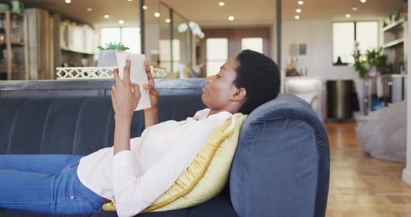 Happy african american woman lying on sofa in living room, reading book