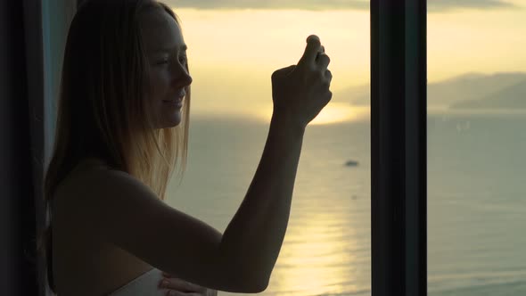 A Young Woman in An Apartment with A Panoramic View on The Coast and Sunrise. She Makes a Photo of A