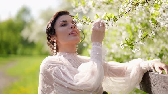 Beautiful Brunette Lady in Dress in Vintage Style is Sniffing Blossoms on Blooming Tree in Spring